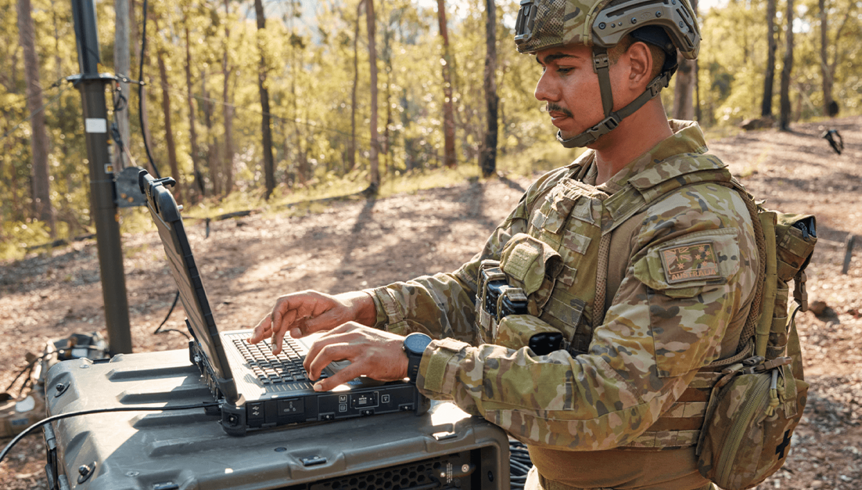 Out in the field, a member of the Army works on his laptop.