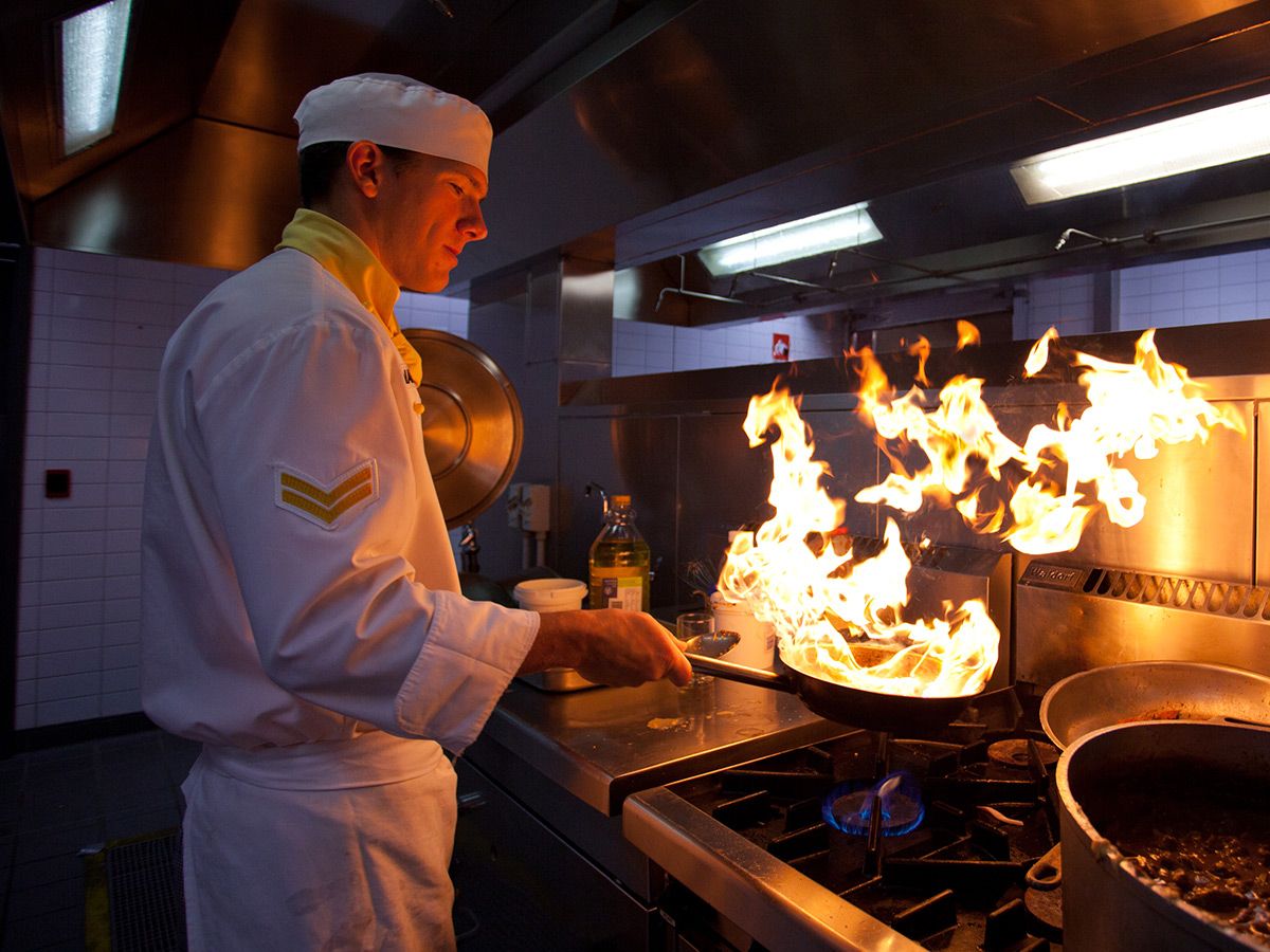 A chef in a kitchen holds a pan over a flaming stove.
