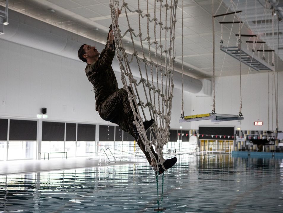 Tim climbs up a net that's strung up over a indoor swimming pool.