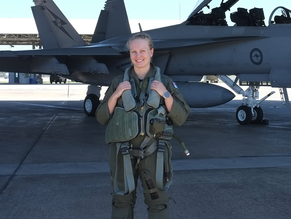 Jasmine is wearing full flying gear, standing in front of a jet smiling.